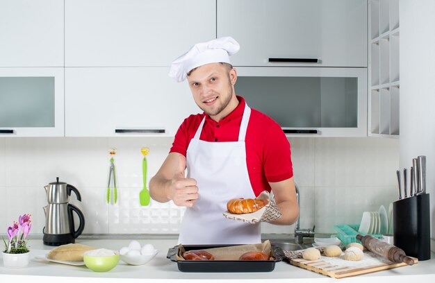 Vorderansicht eines jungen lächelnden männlichen Kochs, der einen Halter mit einem frisch gebackenen Gebäck trägt und in der weißen Küche eine gute Geste macht