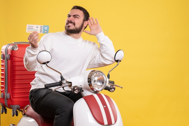 Vorderansicht eines jungen lächelnden reisenden Mannes, der auf einem Motorrad mit einem Koffer darauf sitzt und ein Ticket hält, das dem letzten Tratsch auf isoliertem gelbem Hintergrund zuhört