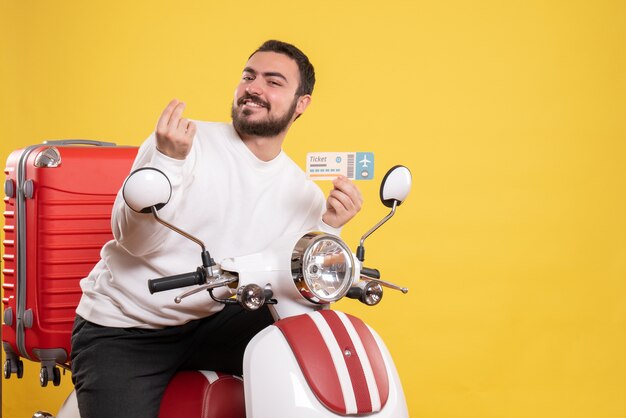 Vorderansicht eines jungen lächelnden reisenden Mannes, der auf einem Motorrad mit einem Koffer darauf sitzt und ein Ticket hält, das Geld auf einem isolierten gelben Hintergrund macht