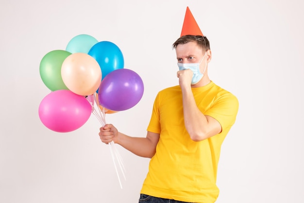 Vorderansicht eines jungen Mannes, der bunte Luftballons in steriler Maske hält und an weißer Wand hustet