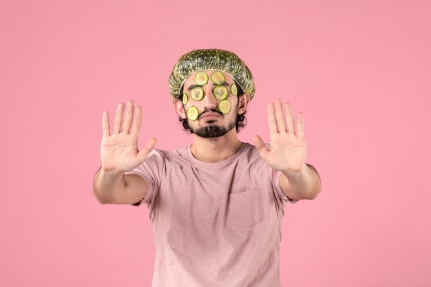 Vorderansicht eines jungen Mannes, der eine Gurkenmaske auf seinem Gesicht an einer rosa Wand anwendet