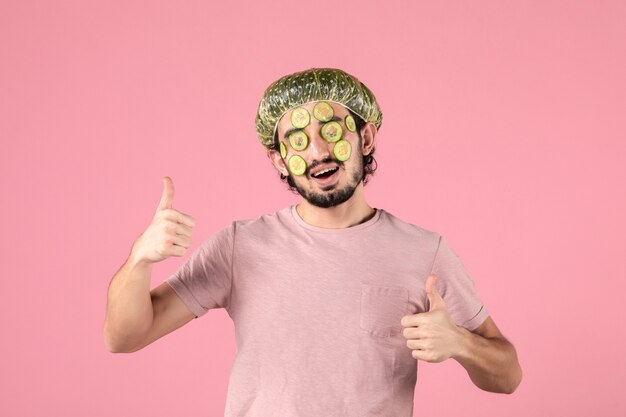 Vorderansicht eines jungen Mannes, der eine Gurkenmaske auf seinem Gesicht an einer rosa Wand anwendet