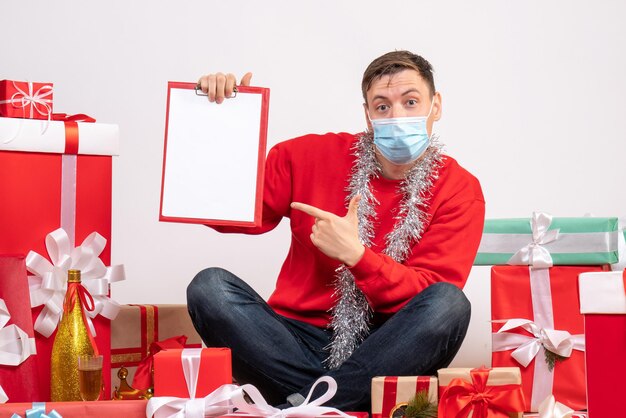 Vorderansicht eines jungen Mannes in Maske, der um Weihnachtsgeschenke sitzt, mit Hinweis auf weißer Wand