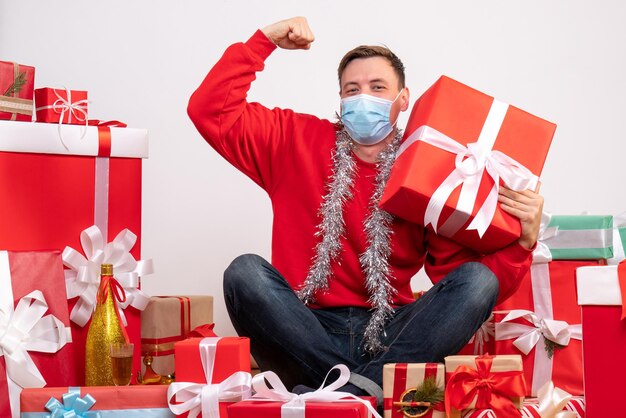 Vorderansicht eines jungen Mannes in steriler Maske, der um Weihnachtsgeschenke auf weißer Wand sitzt