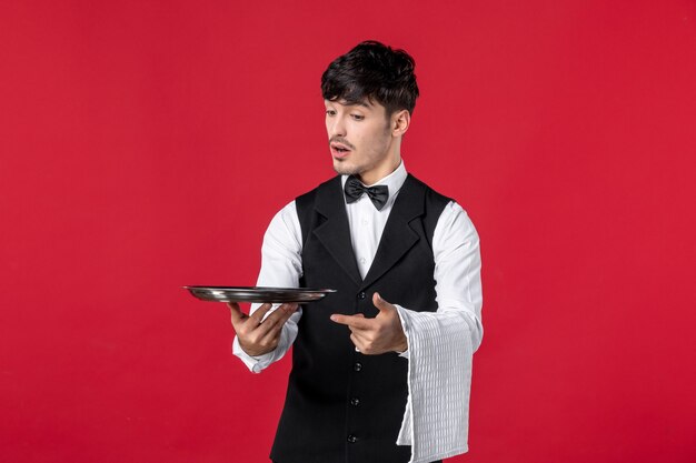 Vorderansicht eines jungen neugierigen männlichen Kellners in Uniform mit Fliege am Hals, der Tablett und Handtuch an roter Wand hält