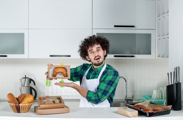 Vorderansicht eines jungen selbstbewussten Mannes, der frisch gebackenes Gebäck in einer kleinen Schachtel in der weißen Küche zeigt