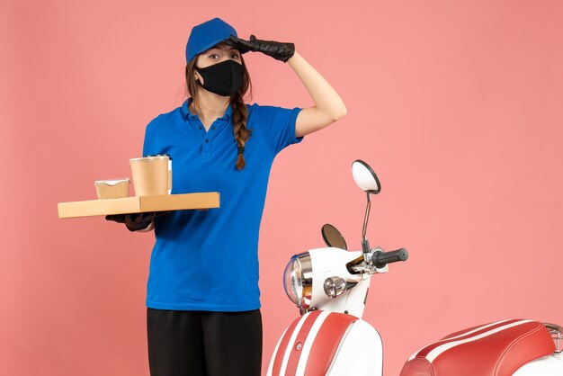Vorderansicht eines Kuriermädchens mit medizinischen Maskenhandschuhen, das neben dem Motorrad steht und kleine Kaffeekuchen hält, die sich auf etwas auf pastellfarbenem Pfirsichhintergrund konzentrieren