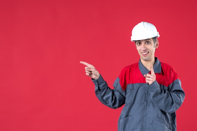 Vorderansicht eines lächelnden jungen Arbeiters in Uniform mit Schutzhelm, der auf etwas zeigt, das einen auf einer isolierten roten Wand zeigt