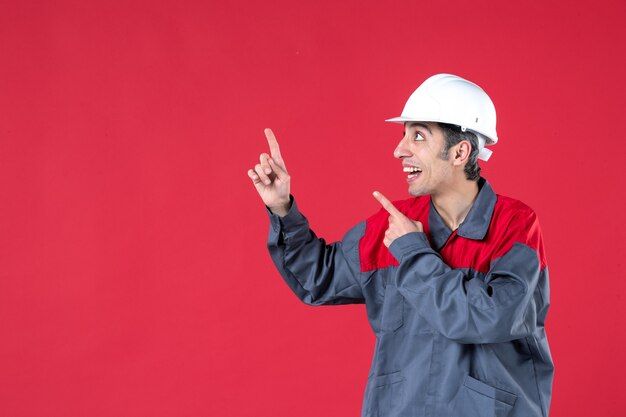 Vorderansicht eines lächelnden jungen Arbeiters in Uniform mit Schutzhelm, der auf isolierte rote Wand zeigt