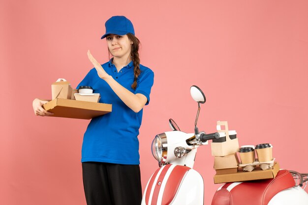 Vorderansicht eines müden Kuriermädchens, das neben dem Motorrad steht und Kaffee und kleine Kuchen auf pastellfarbenem Pfirsichhintergrund hält