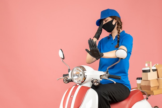 Vorderansicht eines müden Kuriermädchens mit medizinischer Maske und Handschuhen, das auf einem Roller sitzt und Bestellungen auf pastellfarbenem Pfirsichhintergrund liefert