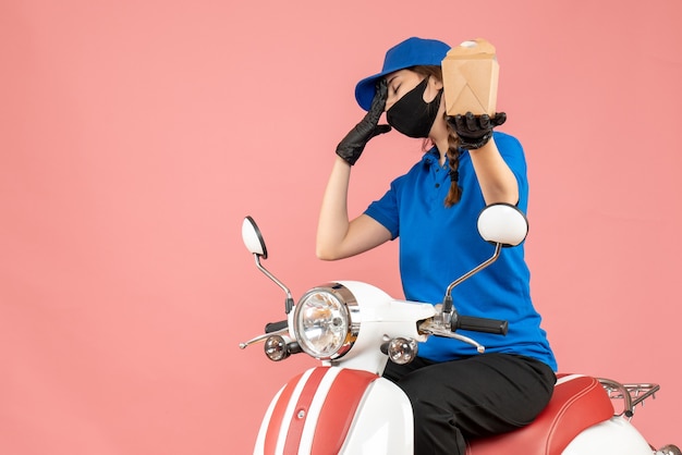 Vorderansicht eines müden Lieferers mit medizinischer Maske und Handschuhen, der auf einem Roller sitzt und Bestellungen auf pastellfarbenem Pfirsichhintergrund liefert