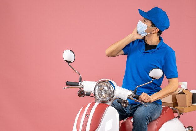 Vorderansicht eines nachdenklichen Kuriermannes in medizinischer Maske mit Hut, der auf einem Roller auf pastellfarbenem Pfirsichhintergrund sitzt