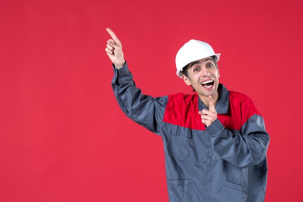 Vorderansicht eines schockierten jungen Arbeiters in Uniform mit Schutzhelm, der auf isolierte rote Wand zeigt