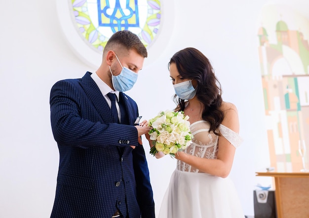 Vorderansicht eines schönen Paares, das Hochzeitskleidung und Schutzmasken auf Gesichtern trägt, die während der standesamtlichen Trauung im Standesamt durch Ringe ausgetauscht werden