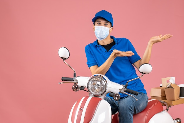 Vorderansicht eines sich wundernden Kuriermannes in medizinischer Maske mit Hut, der auf einem Roller auf pastellfarbenem Pfirsichhintergrund sitzt