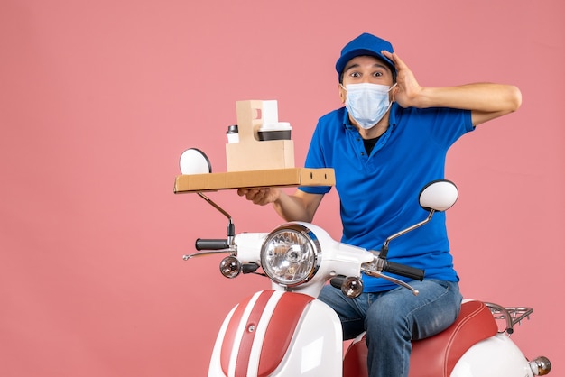 Vorderansicht eines sich wundernden männlichen Lieferers in Maske mit Hut, der auf einem Roller sitzt und Bestellungen auf pastellfarbenem Pfirsichhintergrund liefert