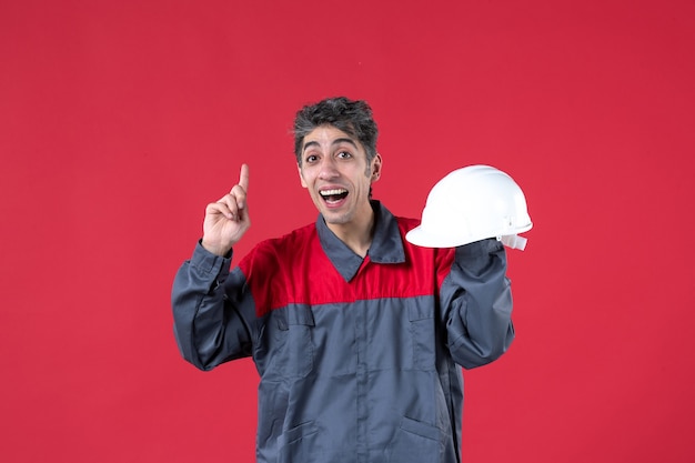 Vorderansicht eines überraschten jungen Arbeiters in Uniform und mit Schutzhelm nach oben auf isolierte rote Wand