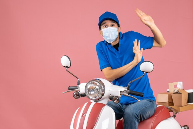 Vorderansicht eines überraschten Lieferers in medizinischer Maske mit Hut, der auf einem Roller auf pastellfarbenem Pfirsichhintergrund sitzt sitting