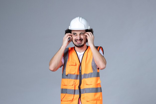 Vorderansicht eines unsicheren, unsicheren männlichen Baumeisters, der Warnweste mit Schutzhelm auf grauer Wellenwand trägt