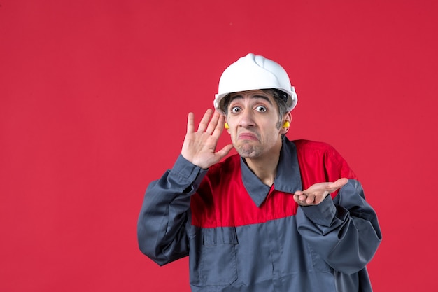Vorderansicht eines verwirrten jungen Arbeiters in Uniform mit Schutzhelm und Ohrstöpseln an isolierter roter Wand