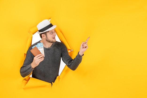 Vorderansicht eines verwirrten jungen Mannes mit einem Hut, der einen ausländischen Pass mit Ticket hält und in einer zerrissenen gelben Wand nach oben zeigt