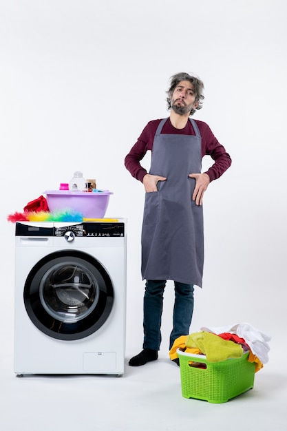 Vorderansicht entschlossener Haushälter, der Hand in die Tasche steckt und neben weißer Waschmaschine auf weißem Hintergrund steht