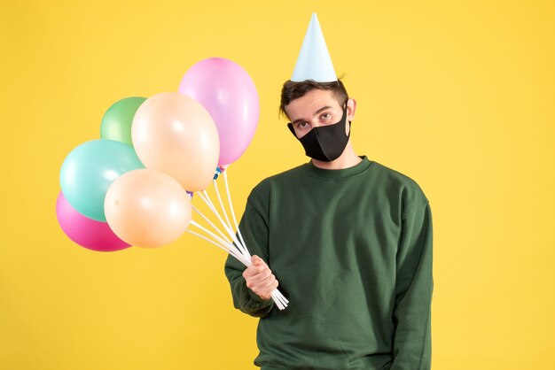 Vorderansicht enttäuschter junger Mann mit Partykappe und bunten Luftballons, die auf Gelb stehen