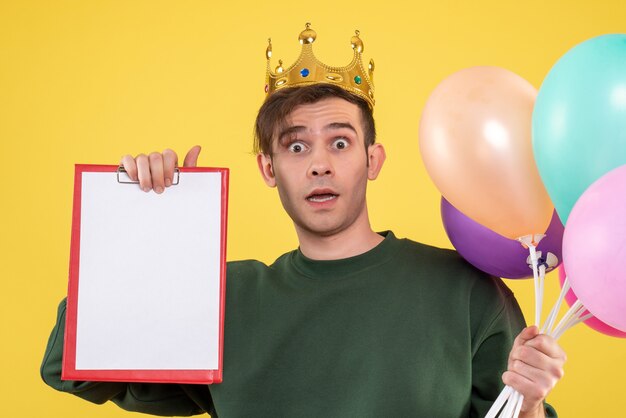 Vorderansicht erstaunte jungen Mann mit Krone, die Luftballons auf Gelb hält