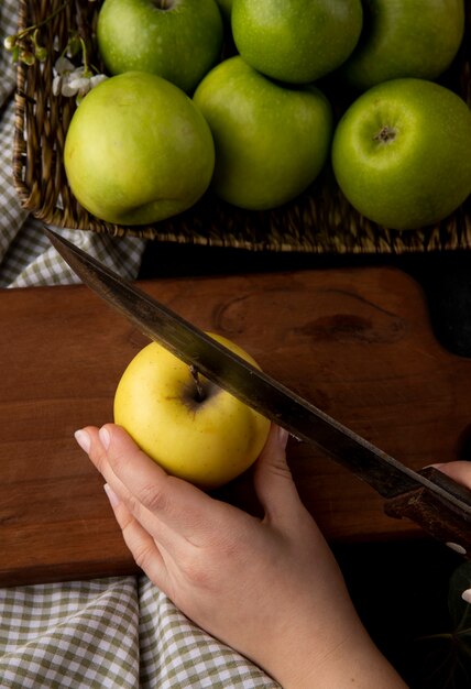 Vorderansicht Frau schneidet gelben Apfel auf einem Brett mit grünen Äpfeln in einem Korb auf einer karierten Tischdecke auf einem Tisch