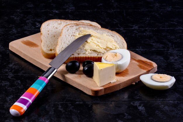 Vorderansicht gekochtes Ei an Bord mit Oliven und Brotscheiben mit Butter und einem Messer