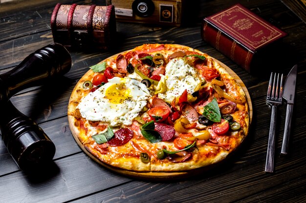 Vorderansicht gemischte Pizza mit Tomaten Oliven Paprika Eier Würstchen auf dem Brett ein Buch ein Messer und eine Gabel auf dem Tisch