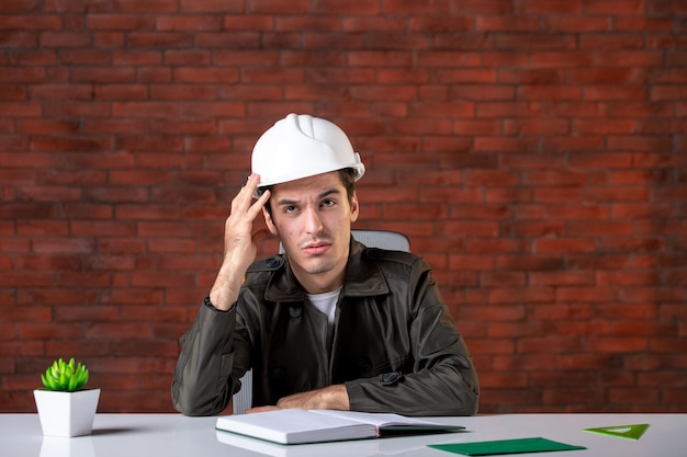 Vorderansicht gestresster männlicher Ingenieur, der hinter seinem Arbeitsplatz sitzt und Notizen schreibt