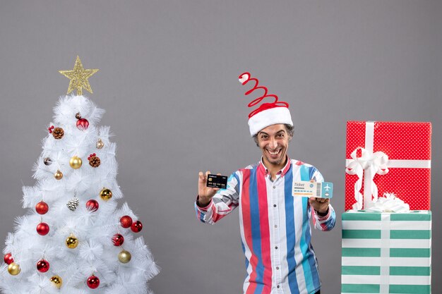 Vorderansicht glücklicher Mann, der Karte und Reiseticket um Weihnachtsbaum hält