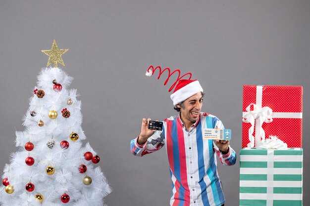 Vorderansicht glücklicher Mann mit blinkendem Auge, das Karte und Reiseticket um Weihnachtsbaum und Geschenke hält