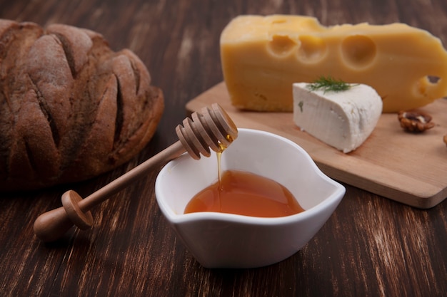 Vorderansicht Honig in einer Untertasse mit einer Vielzahl von Käsesorten auf einem Ständer mit Walnüssen und einem Laib Brot auf einem hölzernen Hintergrund