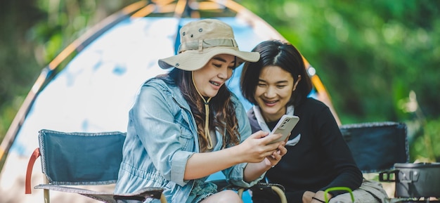 Vorderansicht Junge asiatische hübsche Frau und ihre Freundin, die vor dem Zelt sitzen, benutzen das Handy, um beim Camping im Wald mit Glück zusammen zu fotografieren