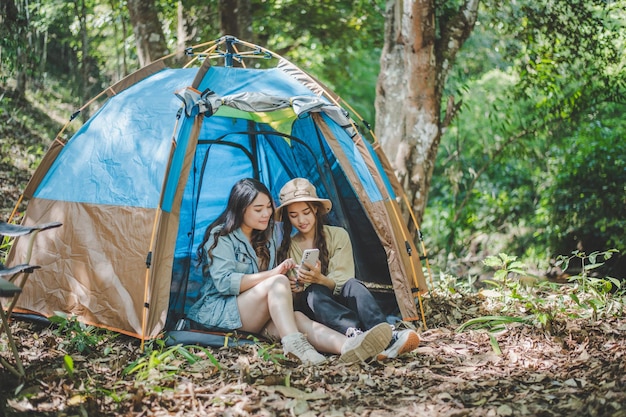 Vorderansicht Junge asiatische hübsche Frau und ihre Freundin, die vor dem Zelt sitzen, benutzen das Handy, um beim Camping im Wald mit Glück zusammen zu fotografieren