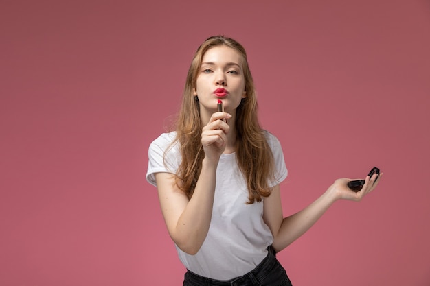 Vorderansicht junge attraktive Frau, die mit Lippenstift und kleinem Spiegel auf der rosa Wandmodellfarbe weiblich jung aufwirft