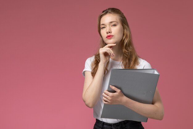 Vorderansicht junge attraktive Frau im weißen T-Shirt, das graues Dokument hält und auf rosa Wandmodell weiblich Pose Farbe weiblich jung denkt