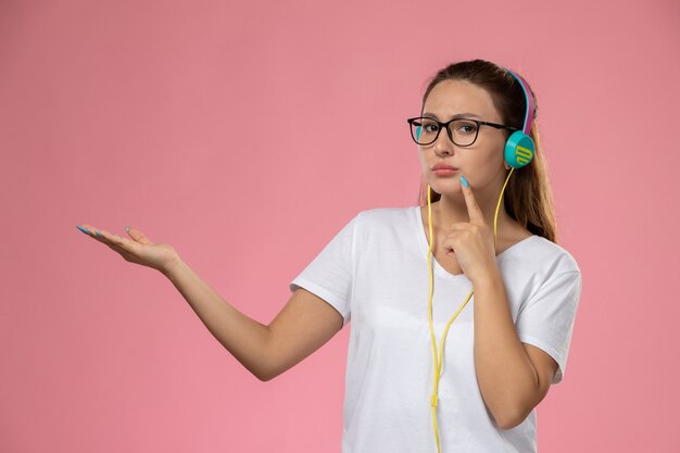 Vorderansicht junge attraktive Frau im weißen T-Shirt, das Musik mit Kopfhörern hört, die auf dem rosa Schreibtisch aufwerfen