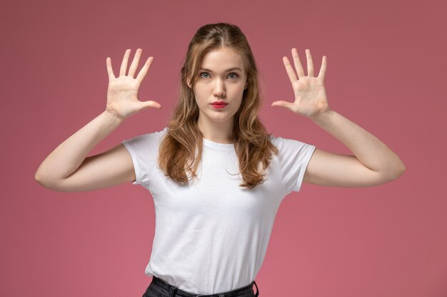 Vorderansicht junge attraktive Frau im weißen T-Shirt mit erhabenen Händen, die auf der rosa Wandmodellfarbe weiblich jung aufwerfen