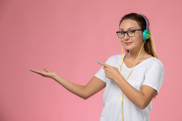 Vorderansicht junge attraktive Frau in weißem T-Shirt, das gerade aufstellt und Musik über Kopfhörer hört, die auf dem rosa Hintergrund lächeln