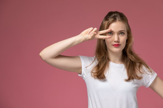 Vorderansicht junge attraktive Frau in weißem T-Shirt, das mit Siegeszeichen auf dem rosa Wandmodell weibliches Posefarbfoto aufwirft