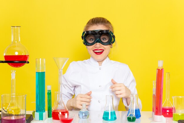 Vorderansicht junge Chemikerin im weißen Anzug mit ed-Lösungen, die mit ihnen arbeiten, die in der Maske auf der gelben Raumchemiewissenschaft sitzen
