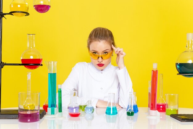 Vorderansicht junge Chemikerin im weißen Anzug vor dem Tisch mit ed-Lösungen, die mit ihnen arbeiten, die auf dem gelben Raumchemie-Wissenschaftsjob verwechseln