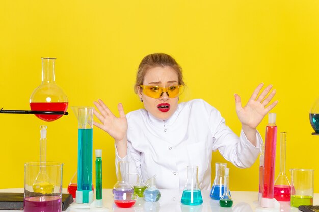 Vorderansicht junge Chemikerin im weißen Anzug vor dem Tisch mit ed-Lösungen, die mit ihnen arbeiten, die auf der Chemie der gelben Raumchemie sitzen