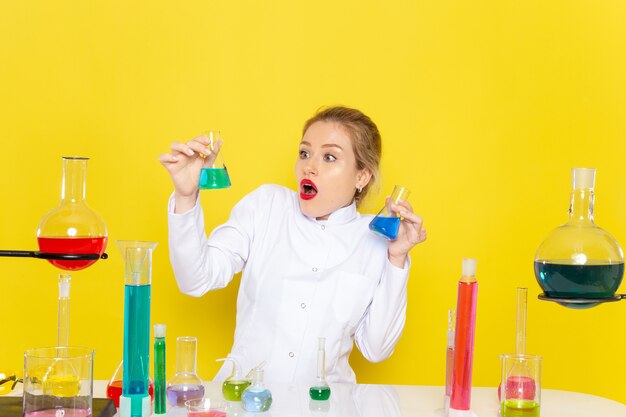 Vorderansicht junge Chemikerin im weißen Anzug vor dem Tisch mit ed-Lösungen, die mit ihnen auf der gelben Raumchemie-Wissenschaft arbeiten