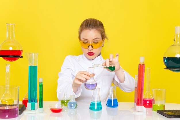 Vorderansicht junge Chemikerin im weißen Anzug vor Tisch mit ed-Lösungen, die mit ihnen arbeiten, die auf der gelben Raumchemie-Wissenschaft verwechseln
