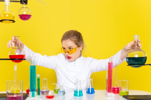Kostenloses Foto vorderansicht junge chemikerin im weißen anzug vor tisch mit ed-lösungen, die mit ihnen arbeiten, die auf der gelben raumchemie-wissenschaft verwechseln